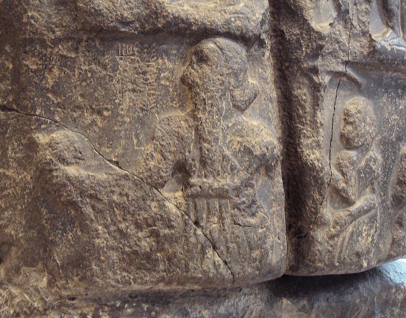 Sargon on his victory stele, with a royal hair bun, holding a mace and wearing a flounced royal coat on his left shoulder with a large belt (left), followed by an attendant holding a royal umbrella.