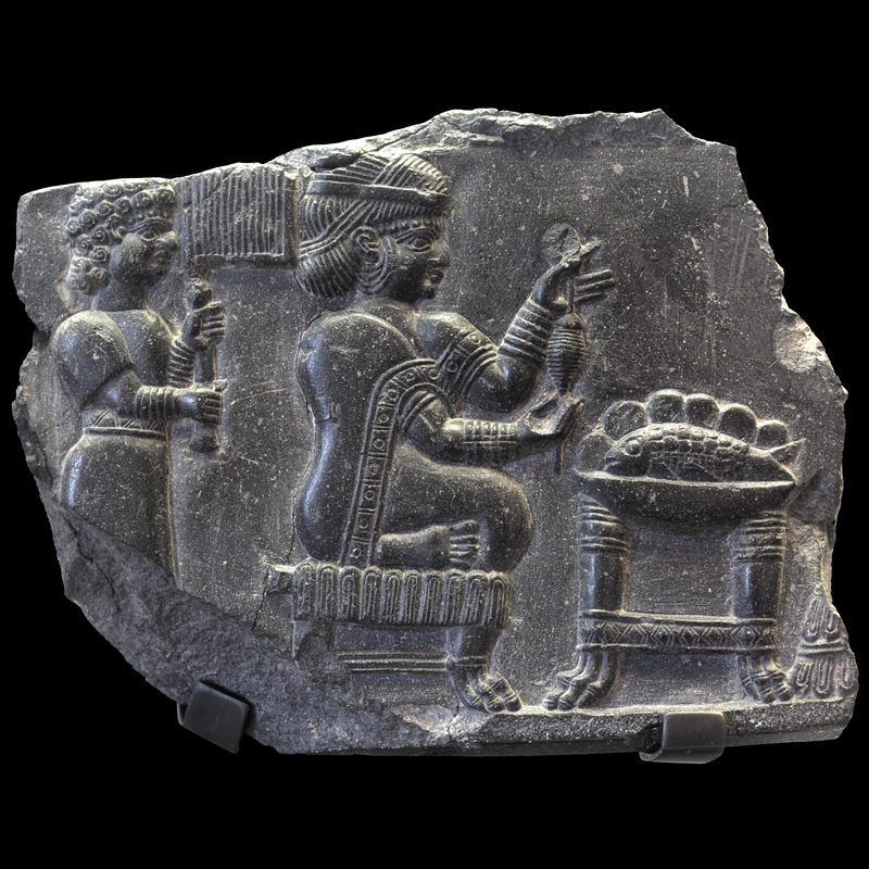 Relief of a woman being fanned by an attendant while she holds what may be a spinning device before a table with a bowl containing a whole fish (700–550 BCE).